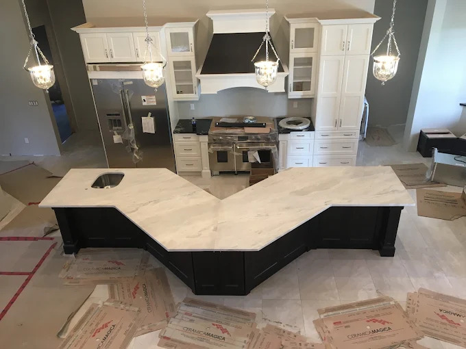 A custom V-shaped island topped in bright white marble with delicate grey veining over a black cabinet base, shown during installation in a white shaker kitchen with a bespoke range hood.